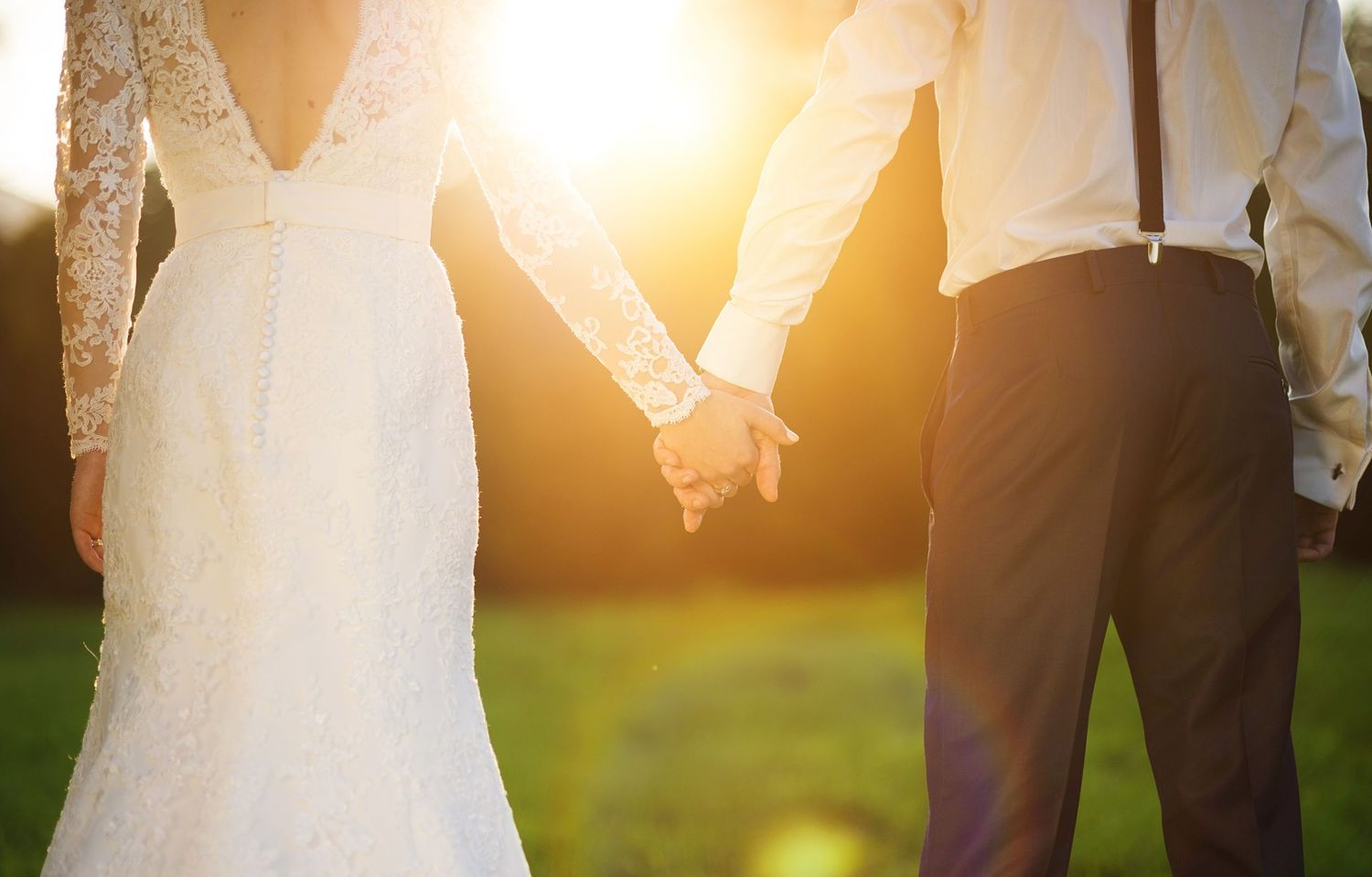 Bride and groom holding hands at sunset in a lush green field, wedding couple backlit by golden light.