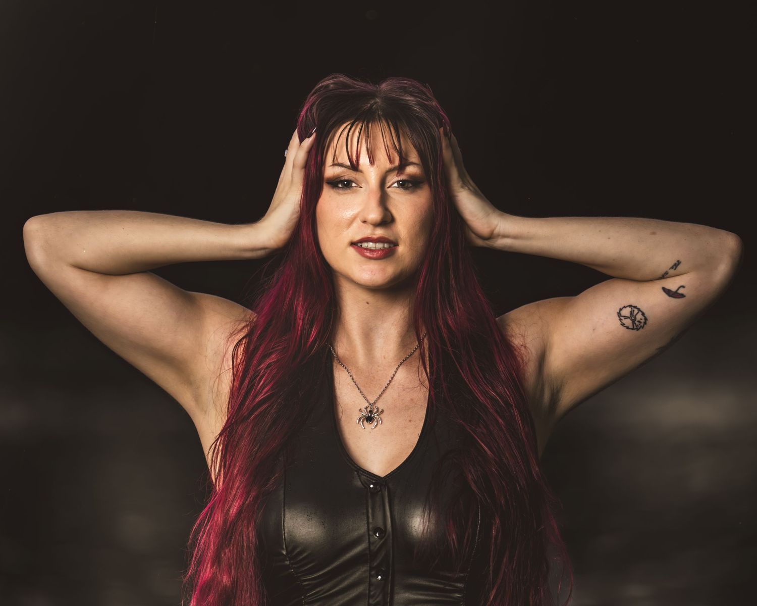Woman with long red hair in black leather top, hands on head, with arm tattoo, against dark background.