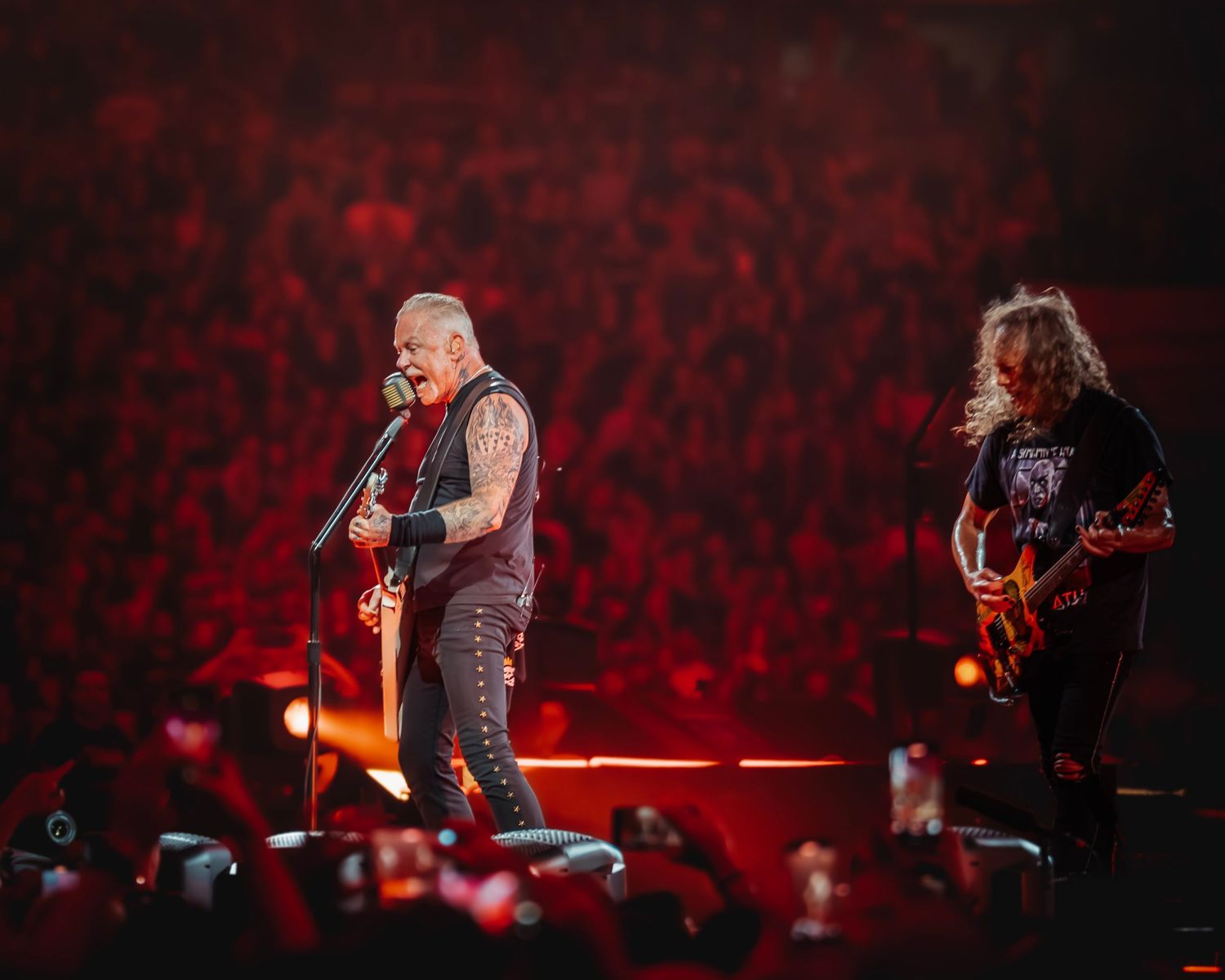Two rock musicians perform on a dramatic red-lit stage before a massive crowd at a live concert.