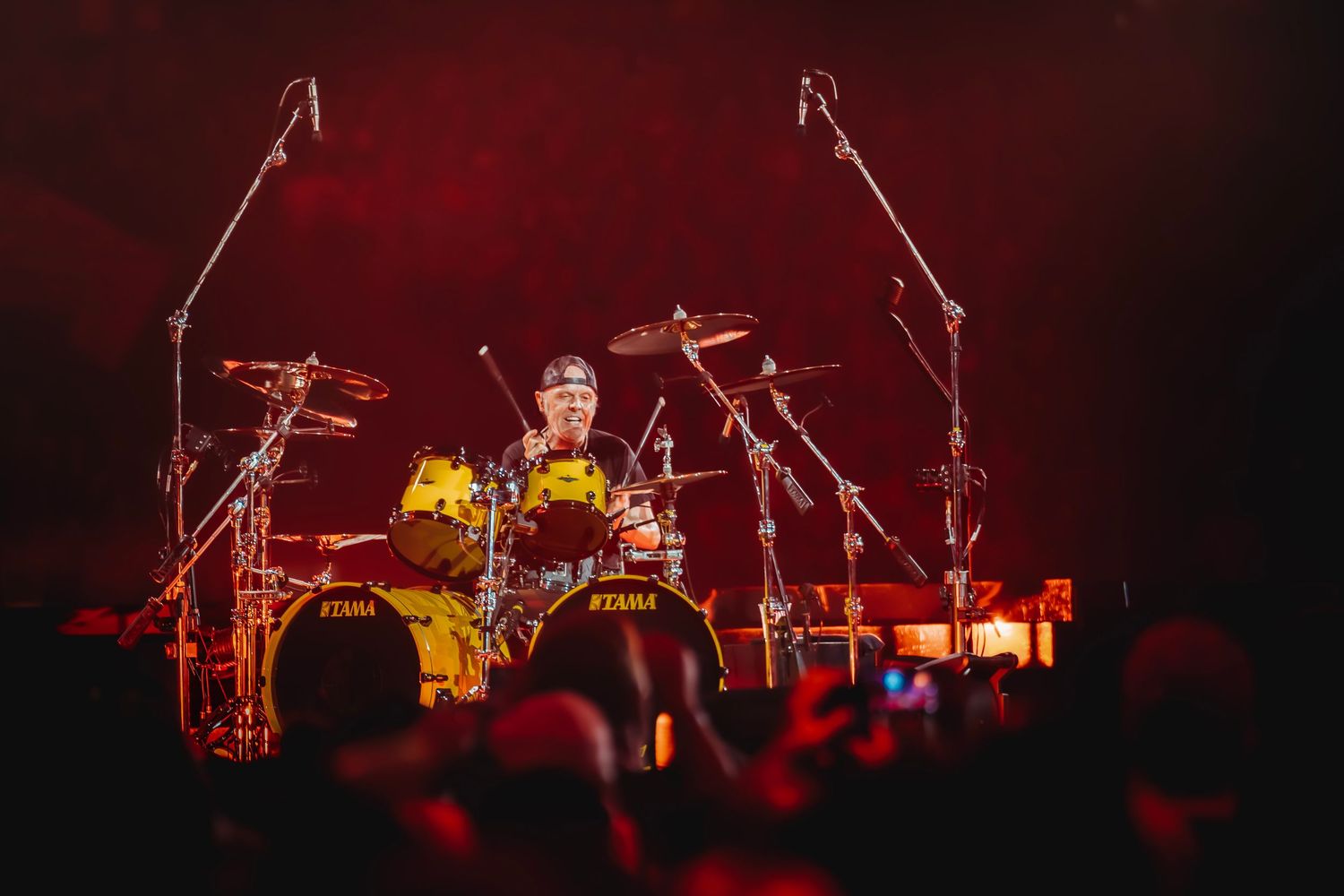Drummer performing on a yellow Tama drum kit under dramatic red stage lighting at a live concert.