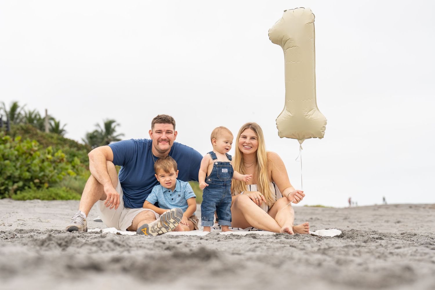 Family of four celebrating baby's first birthday on the beach with a gold number one balloon.