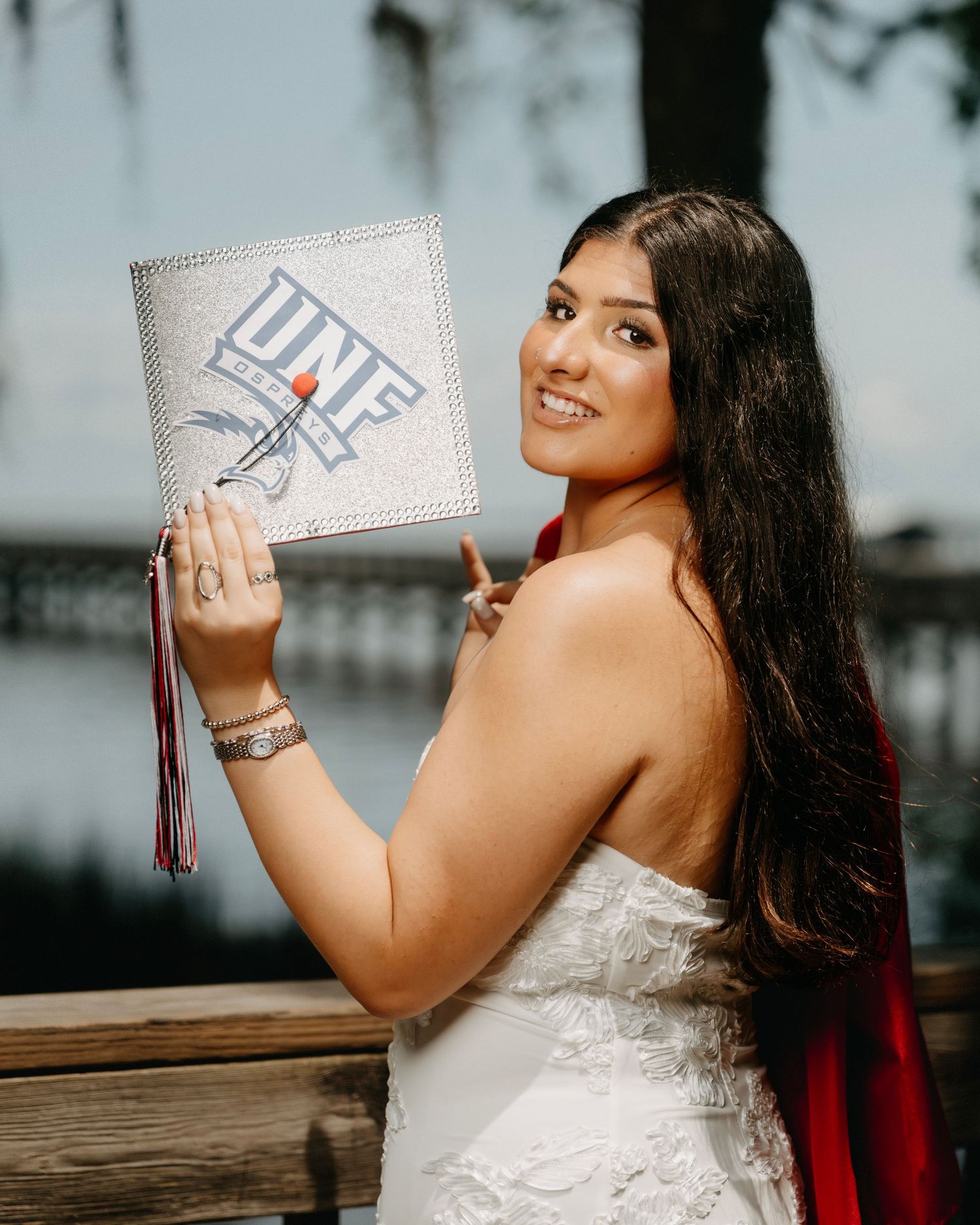 A smiling young woman in a white dress holds up a glittery UNF graduation cap with a red tassel outdoors.