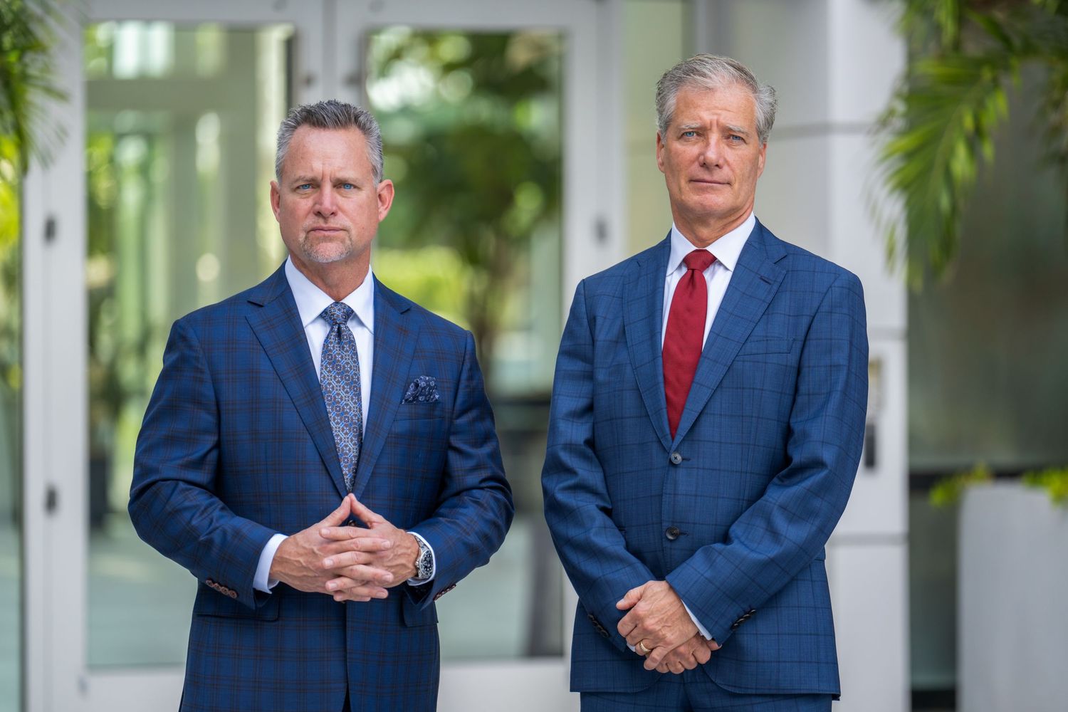 Two professional men in blue suits standing outdoors in a modern building entrance for a formal portrait.