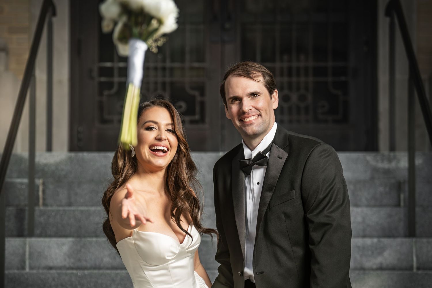 Smiling bride and groom in wedding attire tossing bouquet on church steps.