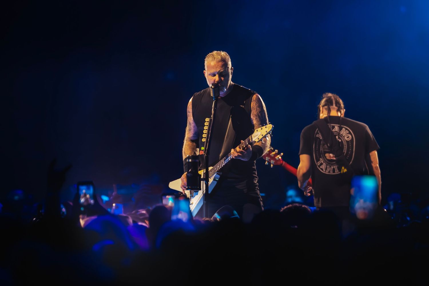 Two rock musicians performing on a dark stage with blue lighting and a crowd in the foreground.