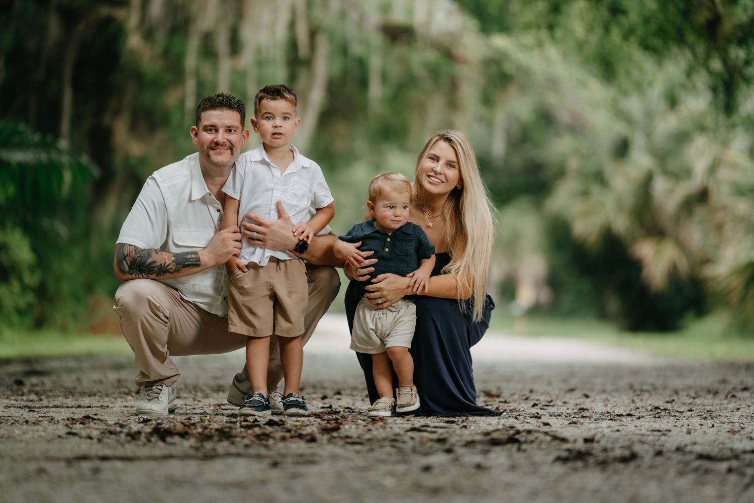 Family of four poses on a scenic tree-lined path, parents crouching with two young boys in a warm outdoor portrait.