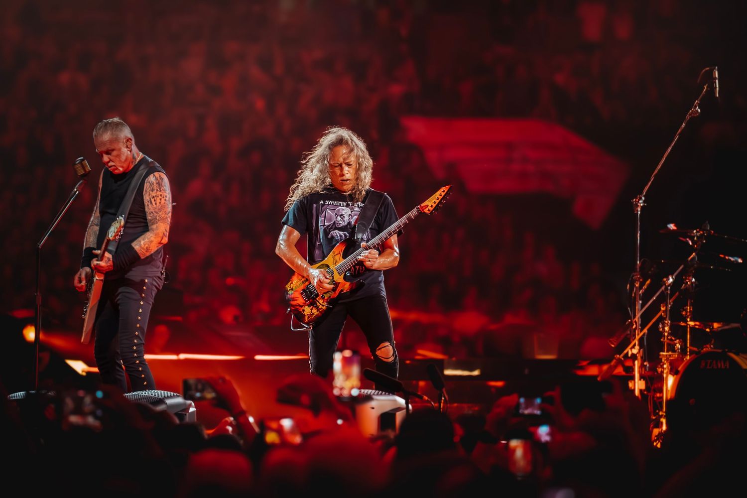 Two rock guitarists performing energetically on stage before a massive crowd under intense red concert lighting.