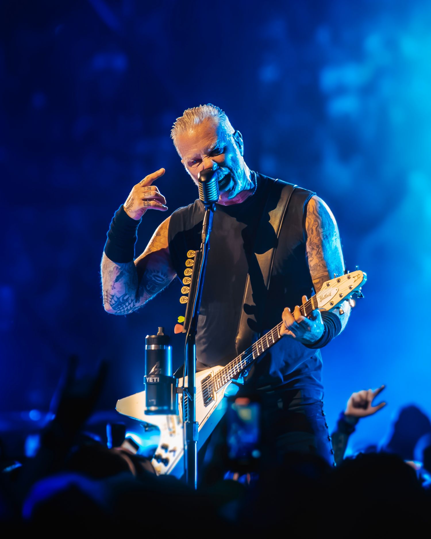 A man with tattoos and a mohawk playing an electric guitar on stage during a concert, with a crowd of people in the background.
