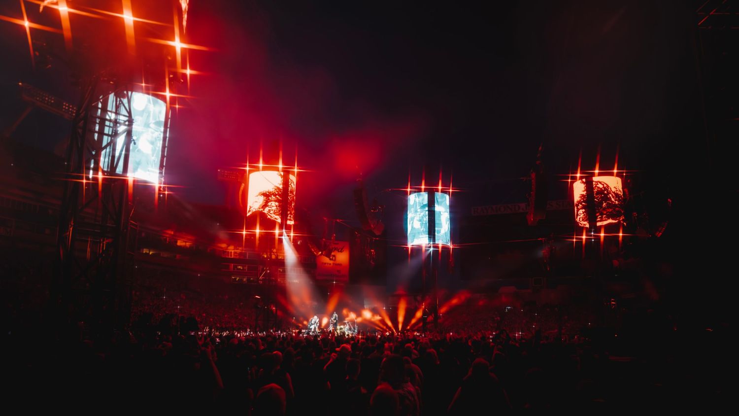 A massive outdoor concert stage lit with dramatic red lights and large video screens above a crowd.