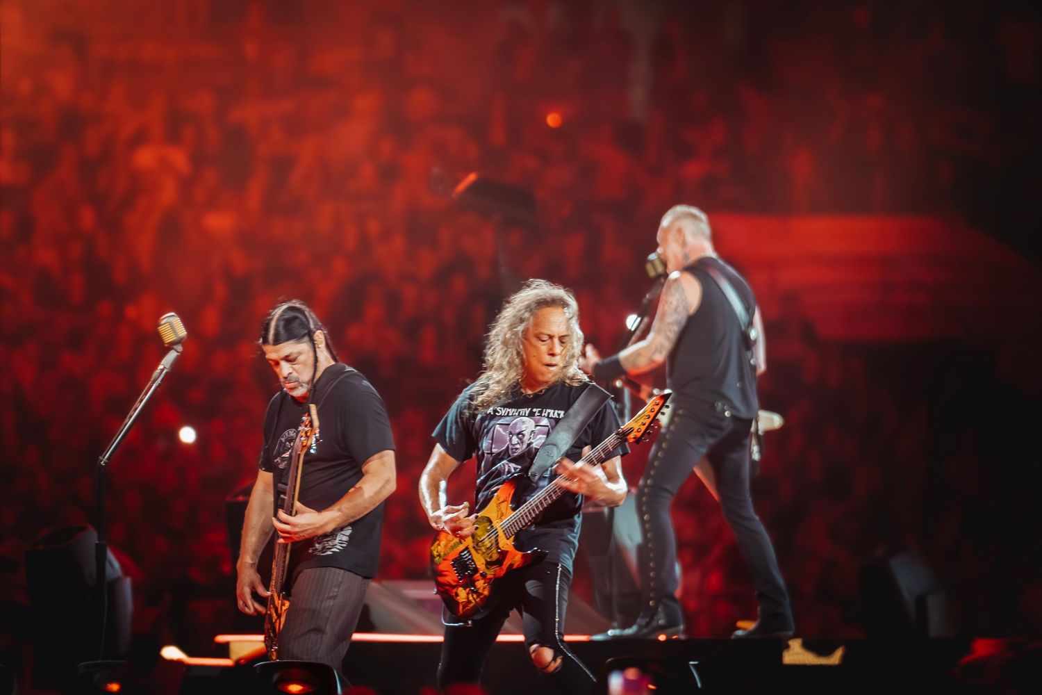 Metallica band members performing live on stage with electric guitars before a massive crowd of fans.