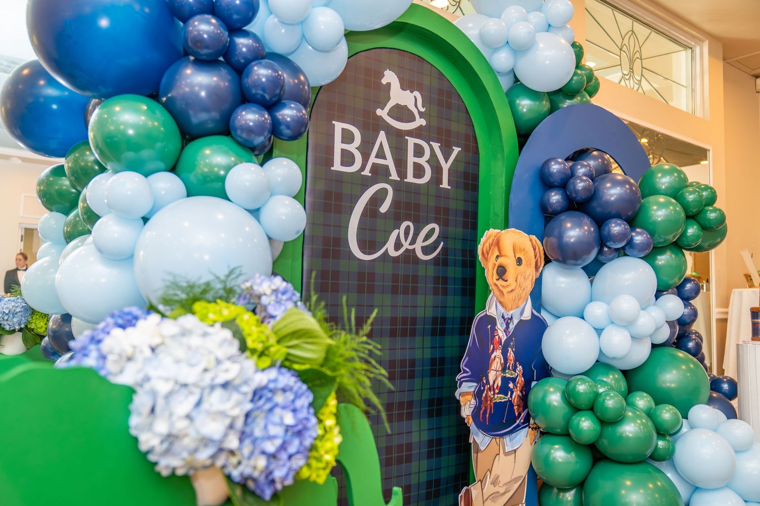 Baby Coe baby shower display with blue and green balloon garland, hydrangeas, and a teddy bear cutout.