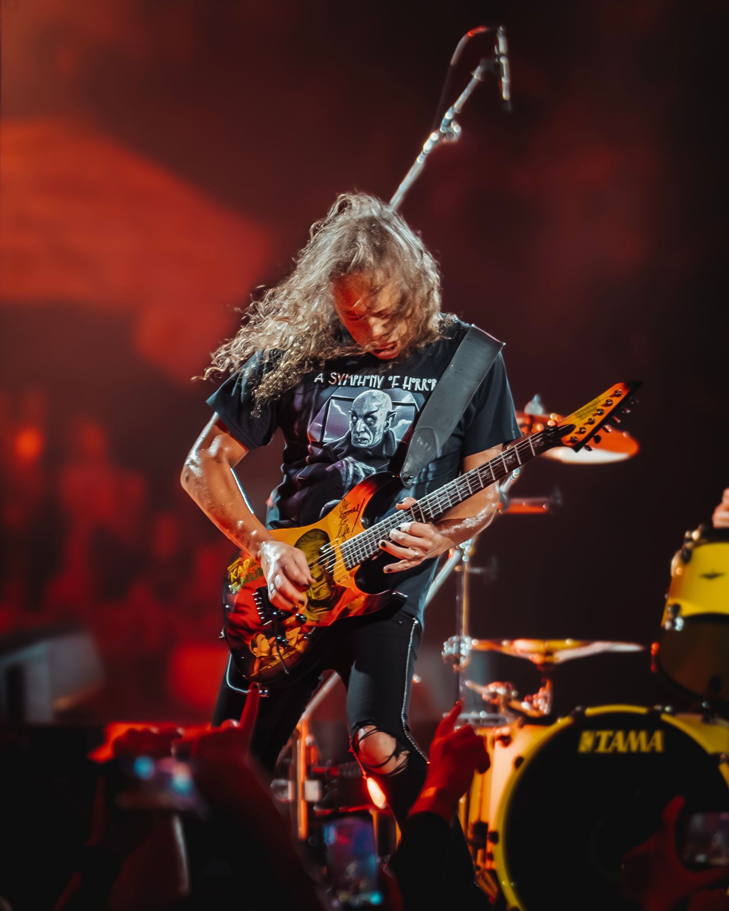 A long-haired guitarist shreds on a colorful electric guitar on a red-lit rock concert stage.