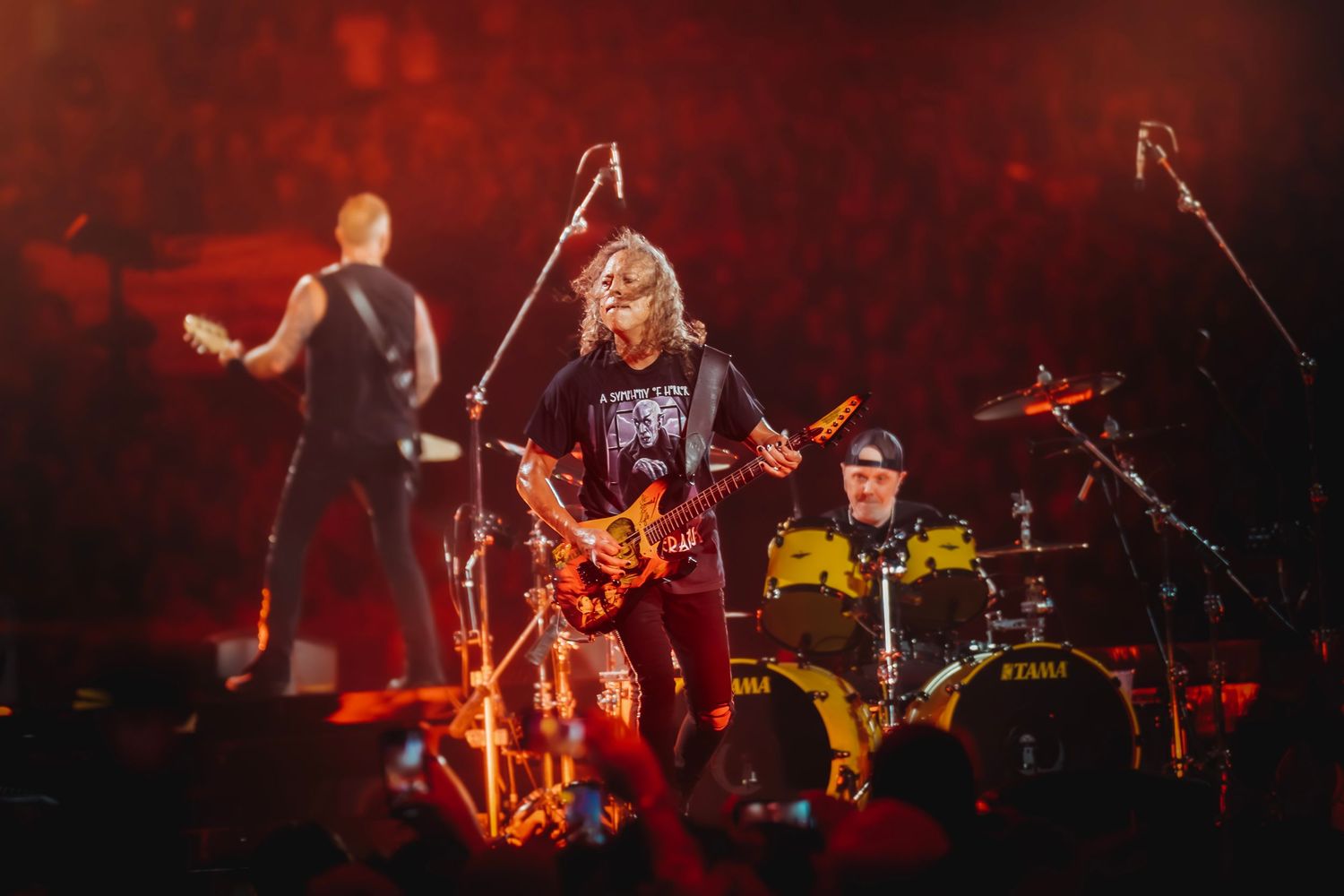 Rock band performing live on stage with guitarist in black tee, bassist, and drummer under dramatic red lighting.