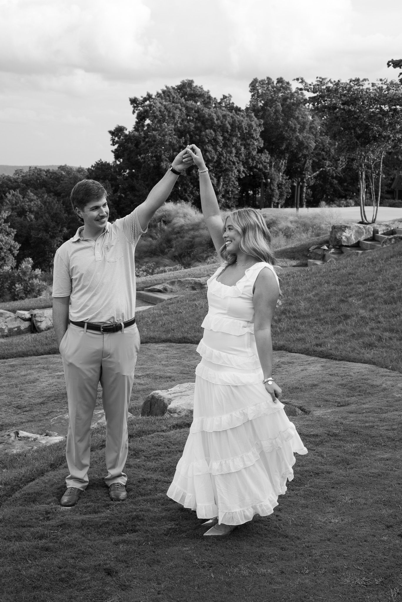 Black and white photo of couple dancing outdoors, man spinning woman in tiered white dress on lawn.