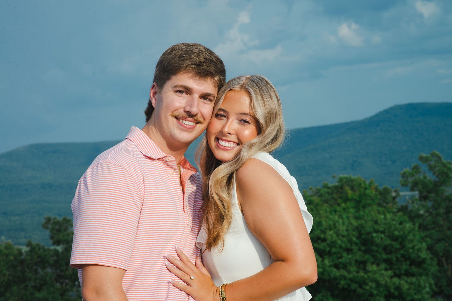 Smiling couple embraces outdoors with mountain and green tree backdrop during engagement photo session.