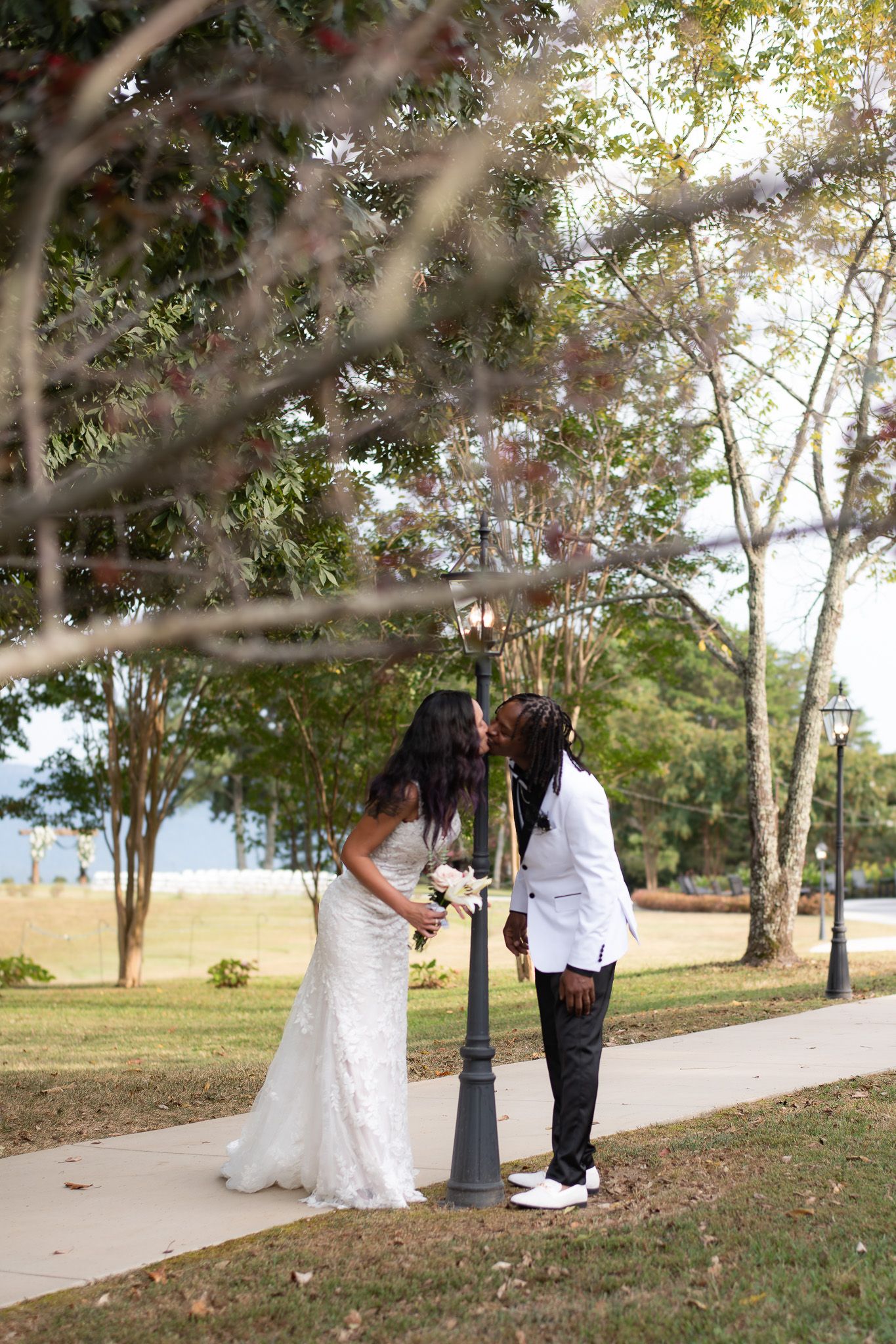 Bride and groom sharing a kiss by a lamppost under blooming trees in a park.