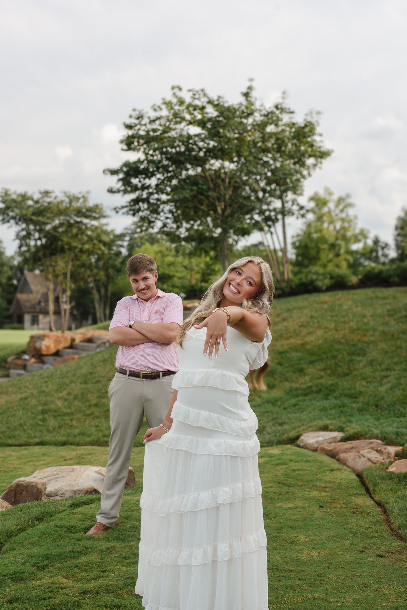 Joyful blonde woman in white ruffled dress showing off engagement ring while fiance stands behind her outdoors.