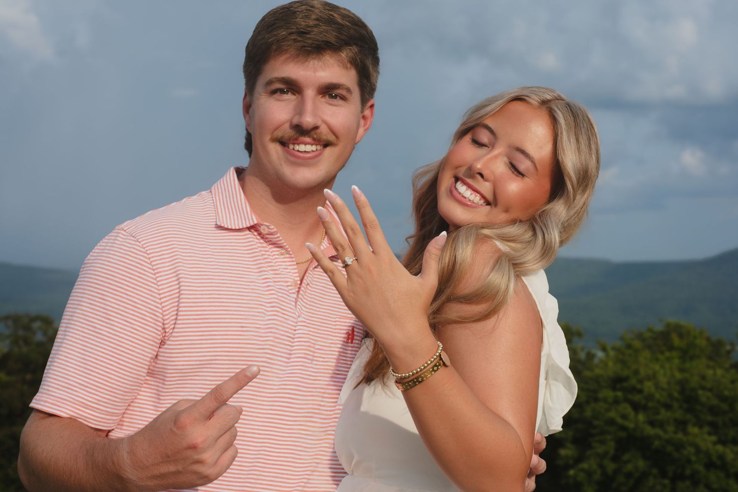 Excited couple shows off engagement ring outdoors with mountains in the background.