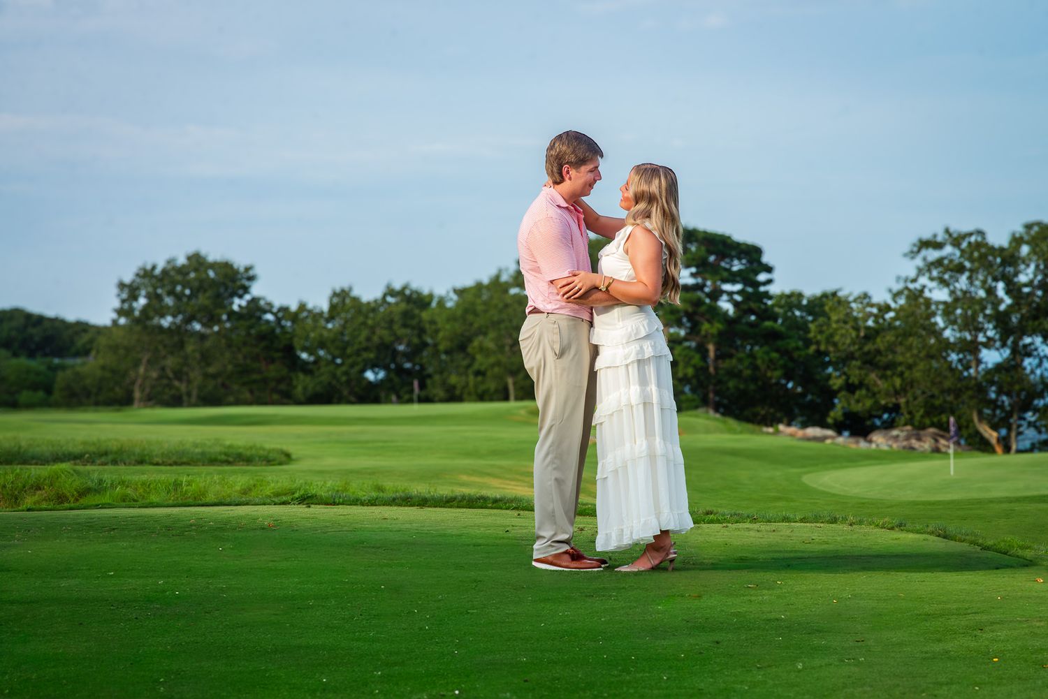 Couple stands together on a golf course green during golden hour engagement photo session.