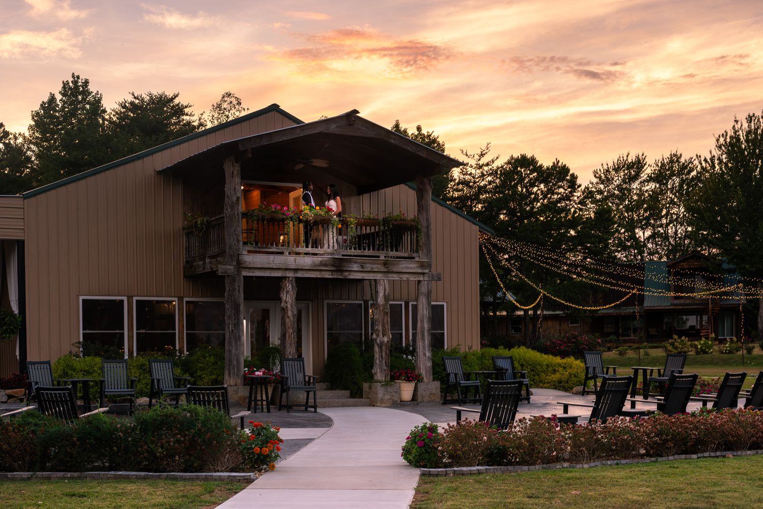 Rustic barn wedding venue at sunset with string lights and guests on upper deck.