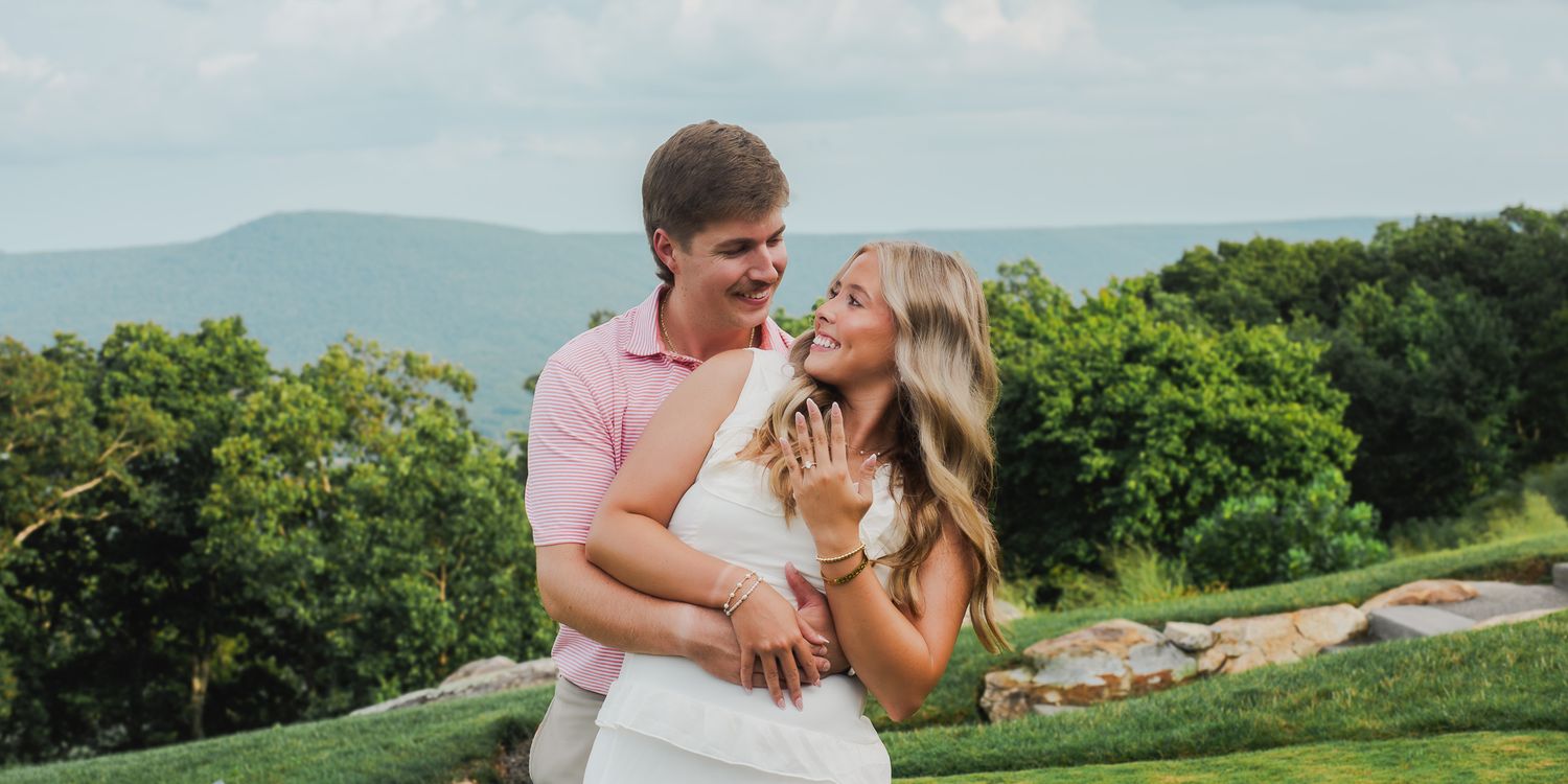 Happy couple laughs and embraces on a lush green lawn with scenic mountain views behind them.
