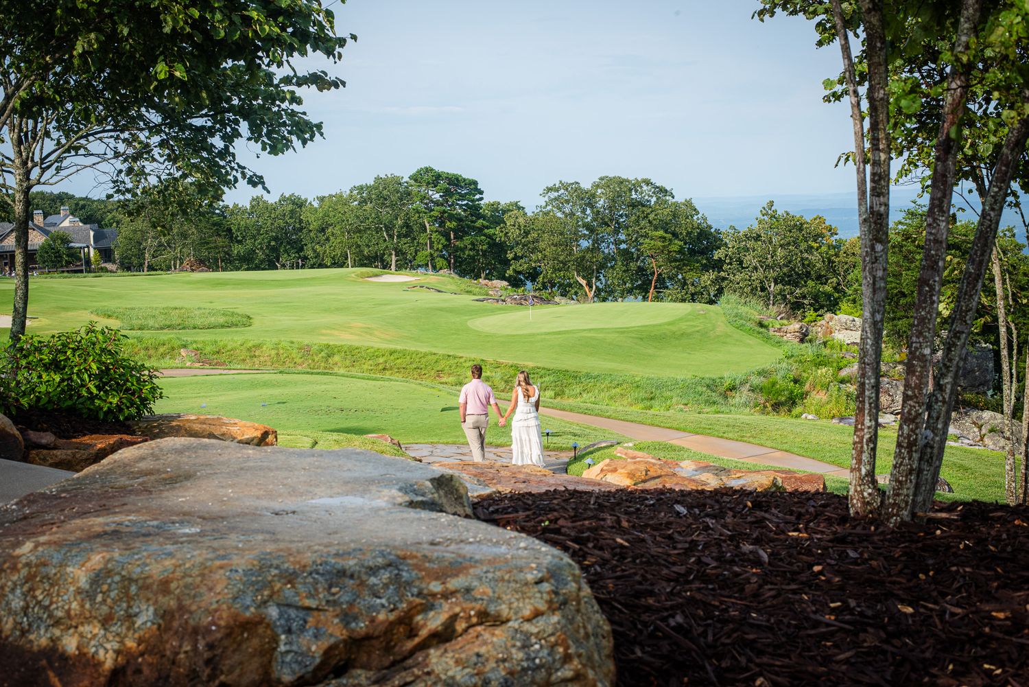 Engaged couple holding hands walking toward lush green golf course framed by trees and rocks.