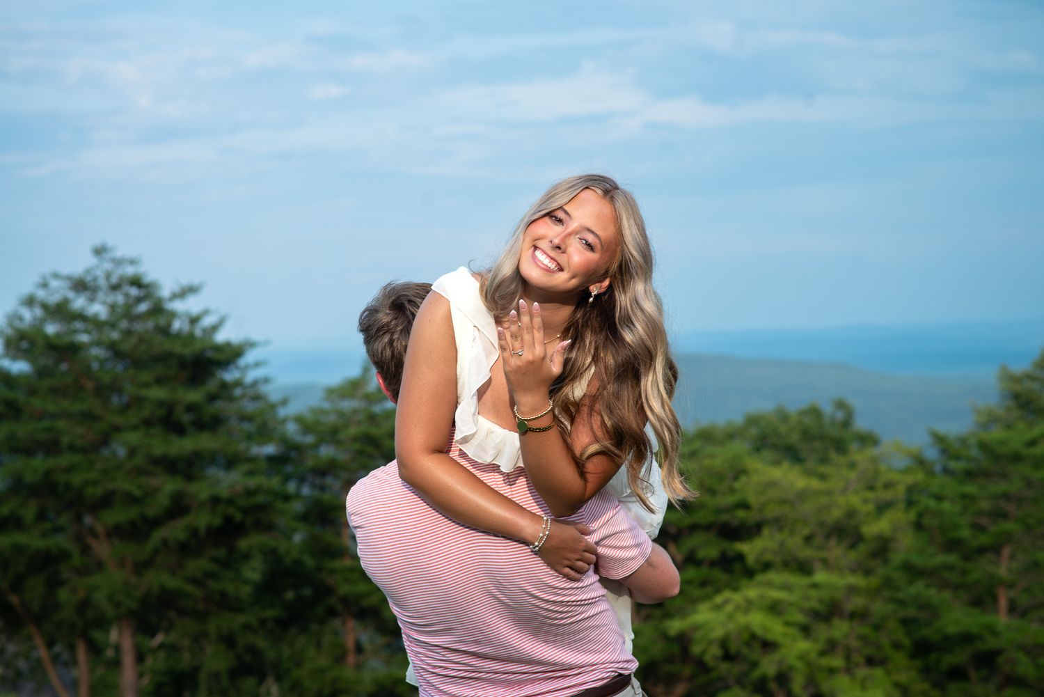Joyful woman showing engagement ring while being lifted by partner with scenic mountain backdrop.