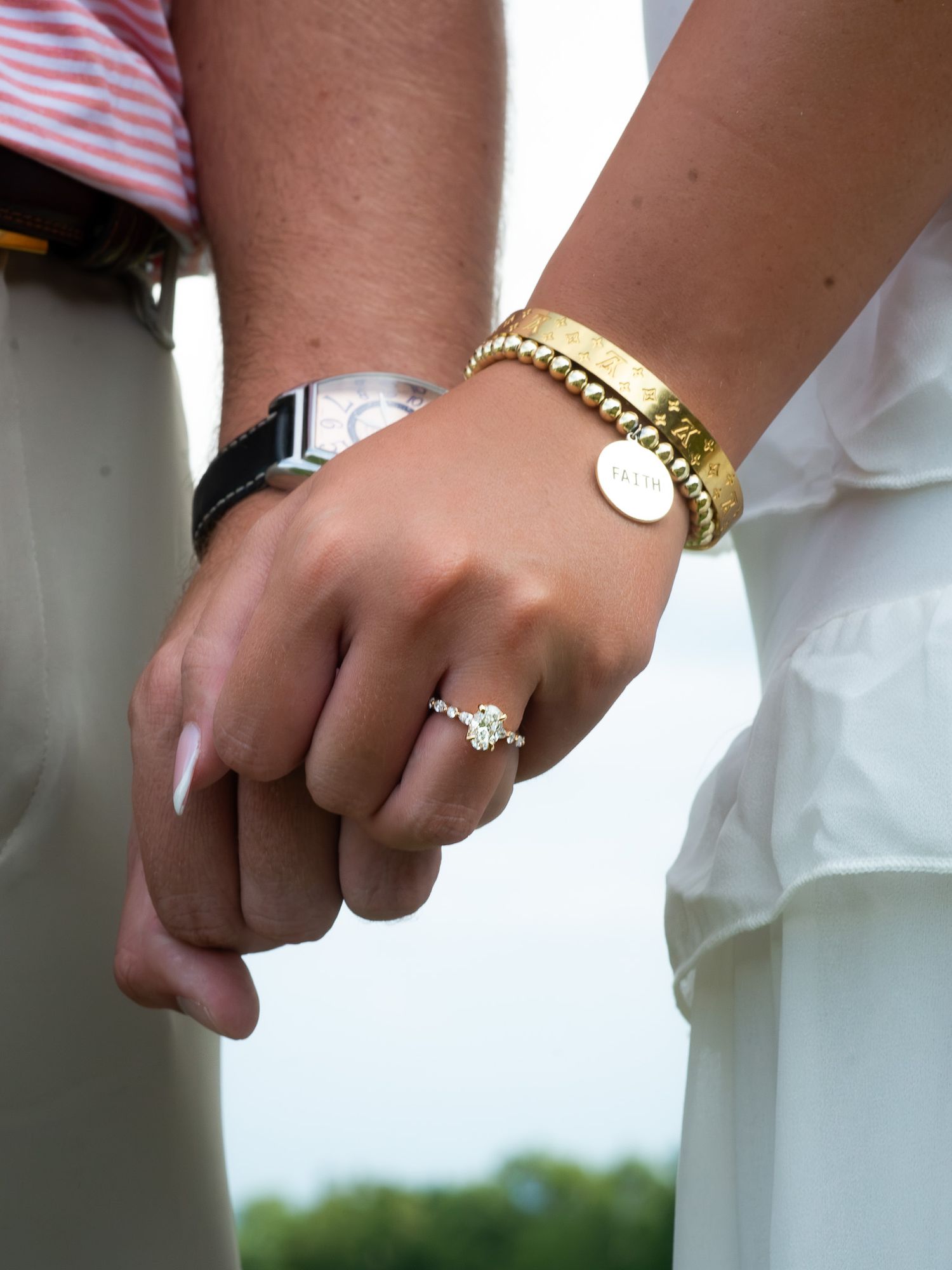 Close-up of engaged couple holding hands showing diamond engagement ring and gold bracelets outdoors.