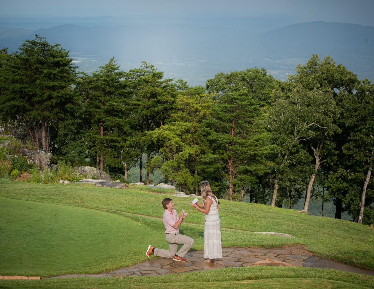 Man proposing on one knee to woman on scenic green golf course with mountain views in background.