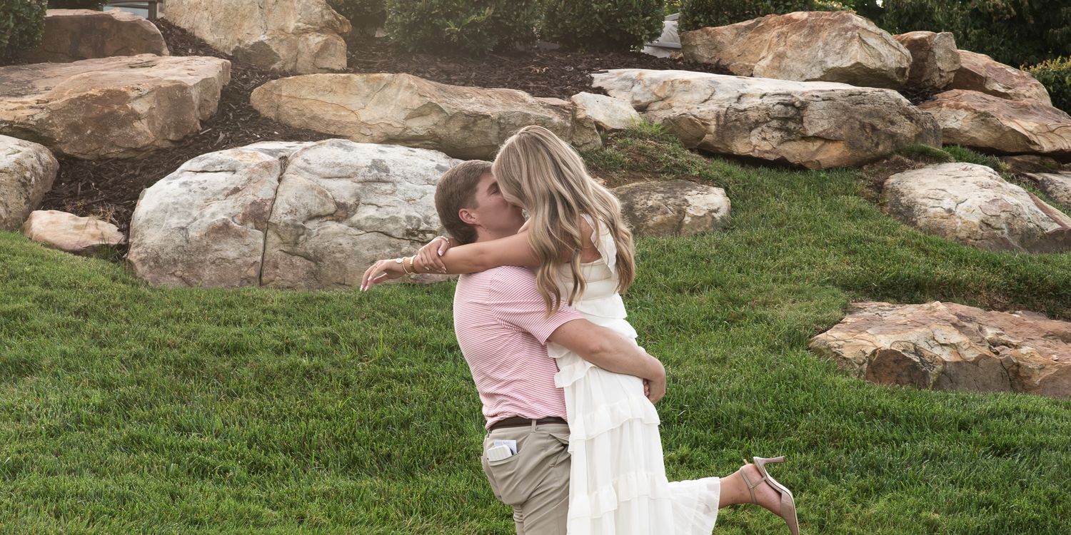 Groom lifts bride in a joyful embrace on a green lawn with large boulders nearby.
