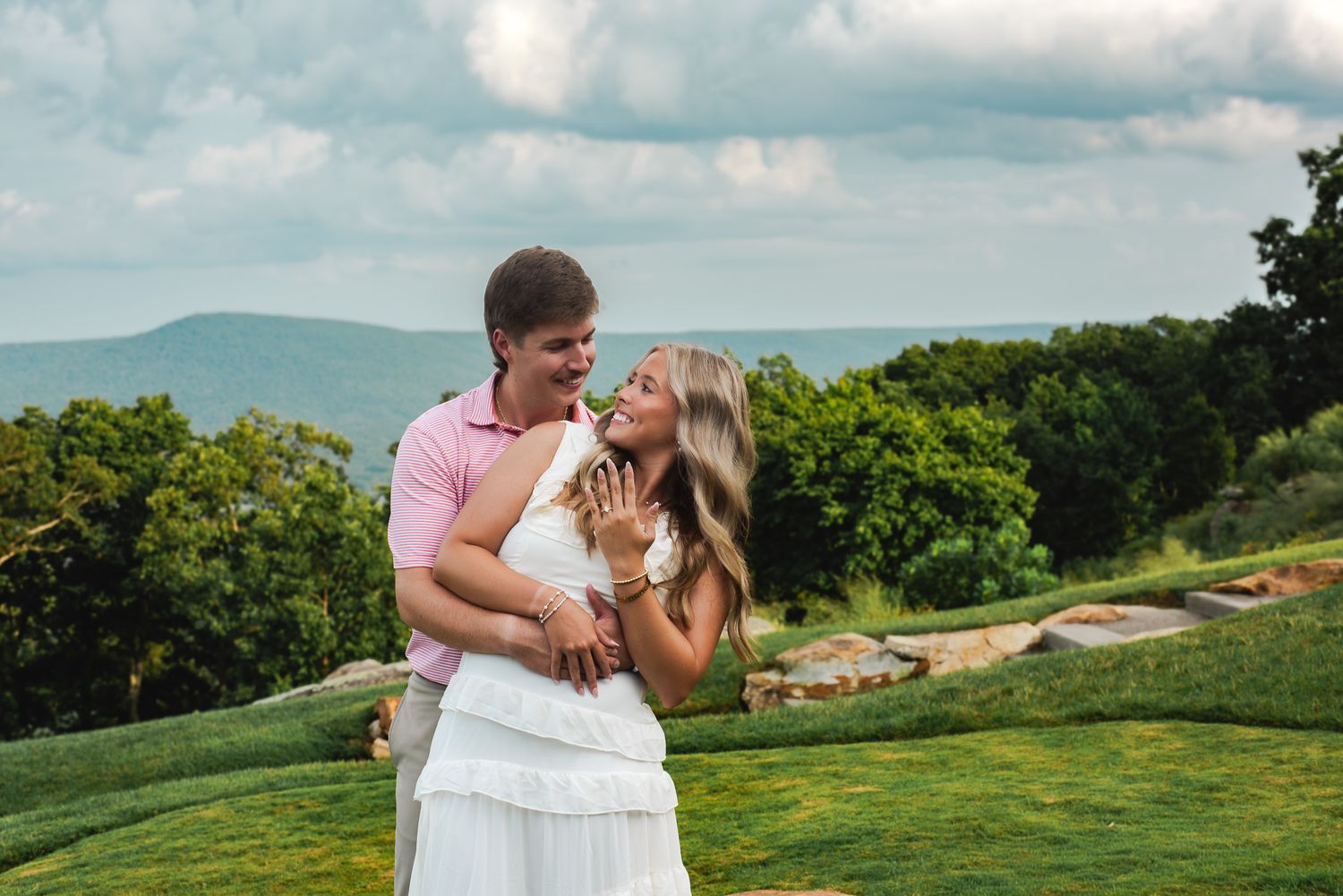 Happy engaged couple embracing and laughing together with scenic green mountain landscape in the background.