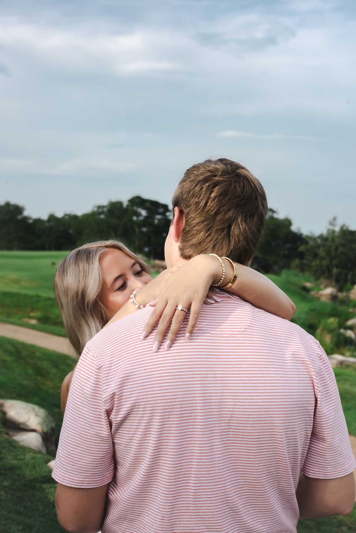Bride embraces groom from behind showing off engagement ring on a scenic golf course.