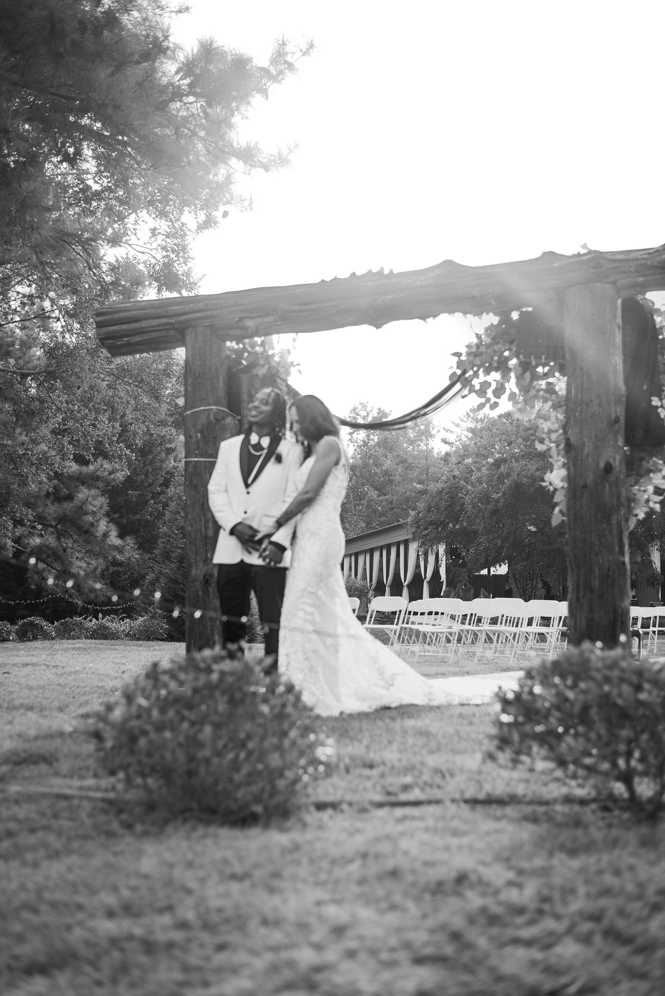 Black and white photo of bride and groom exchanging vows under a rustic wooden arch outdoors.