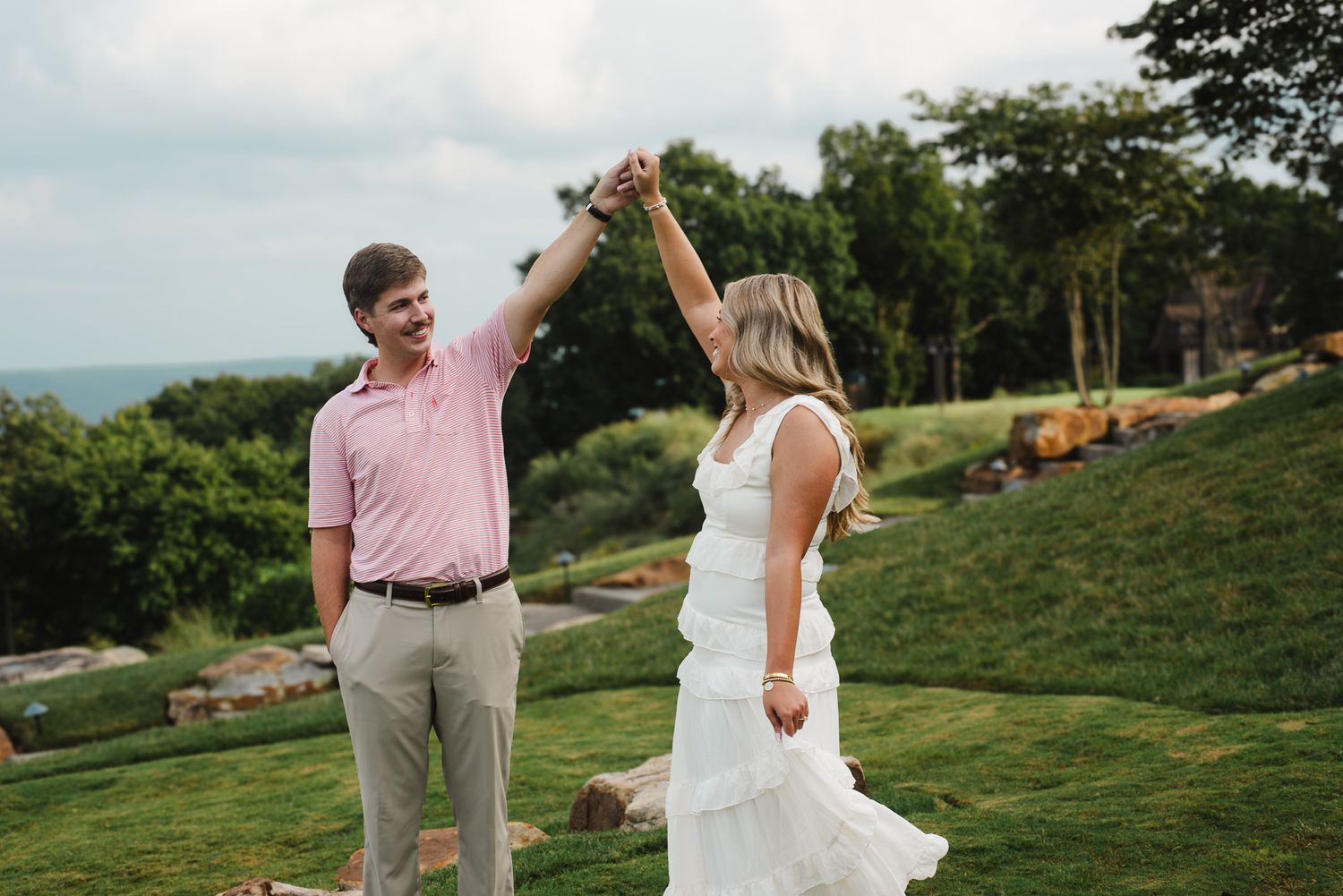 Couple dancing outdoors on green hillside, man in pink polo twirling woman in white dress with mountain views.