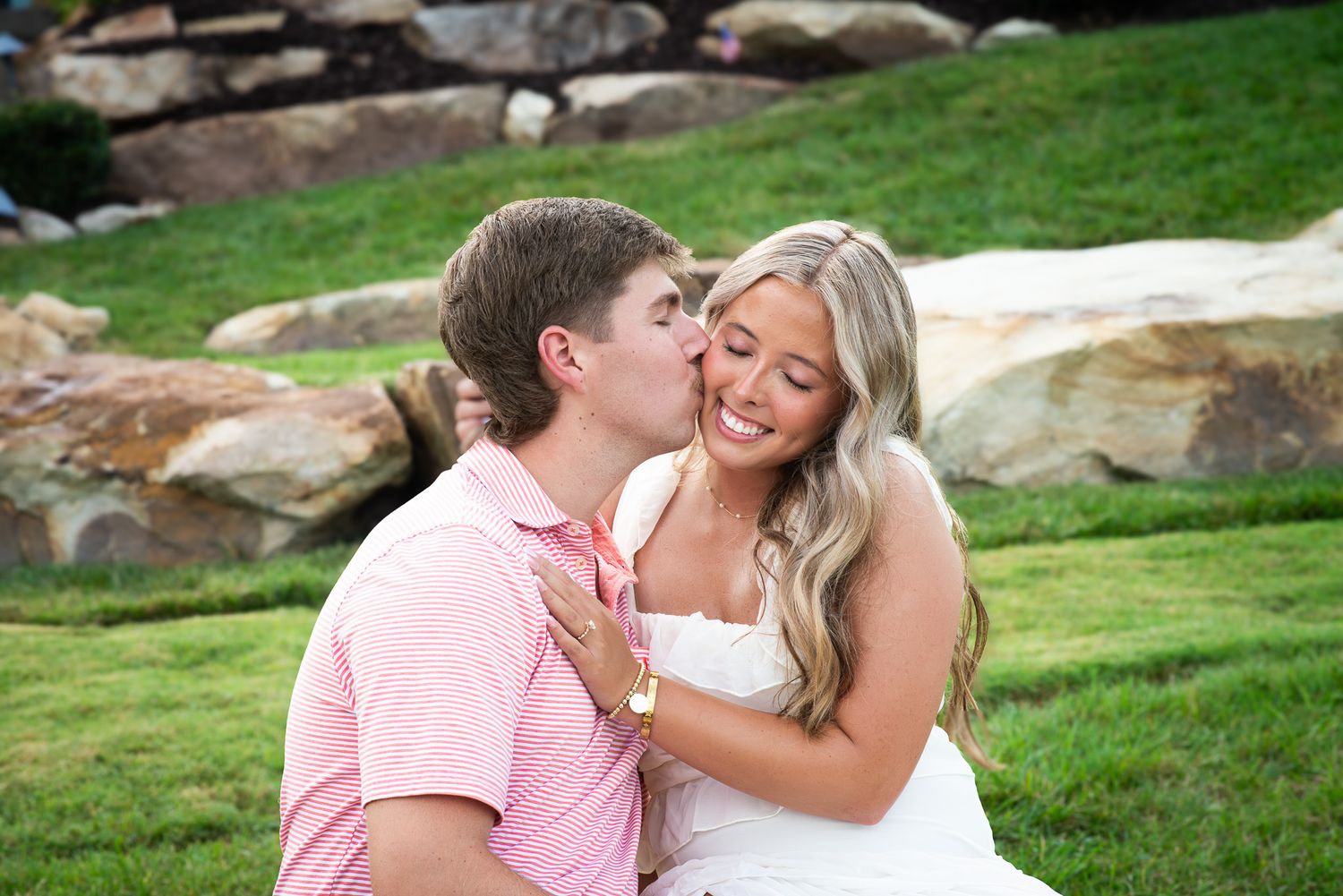 Happy engaged couple sitting on grass, man in pink polo kissing smiling blonde woman in white dress.