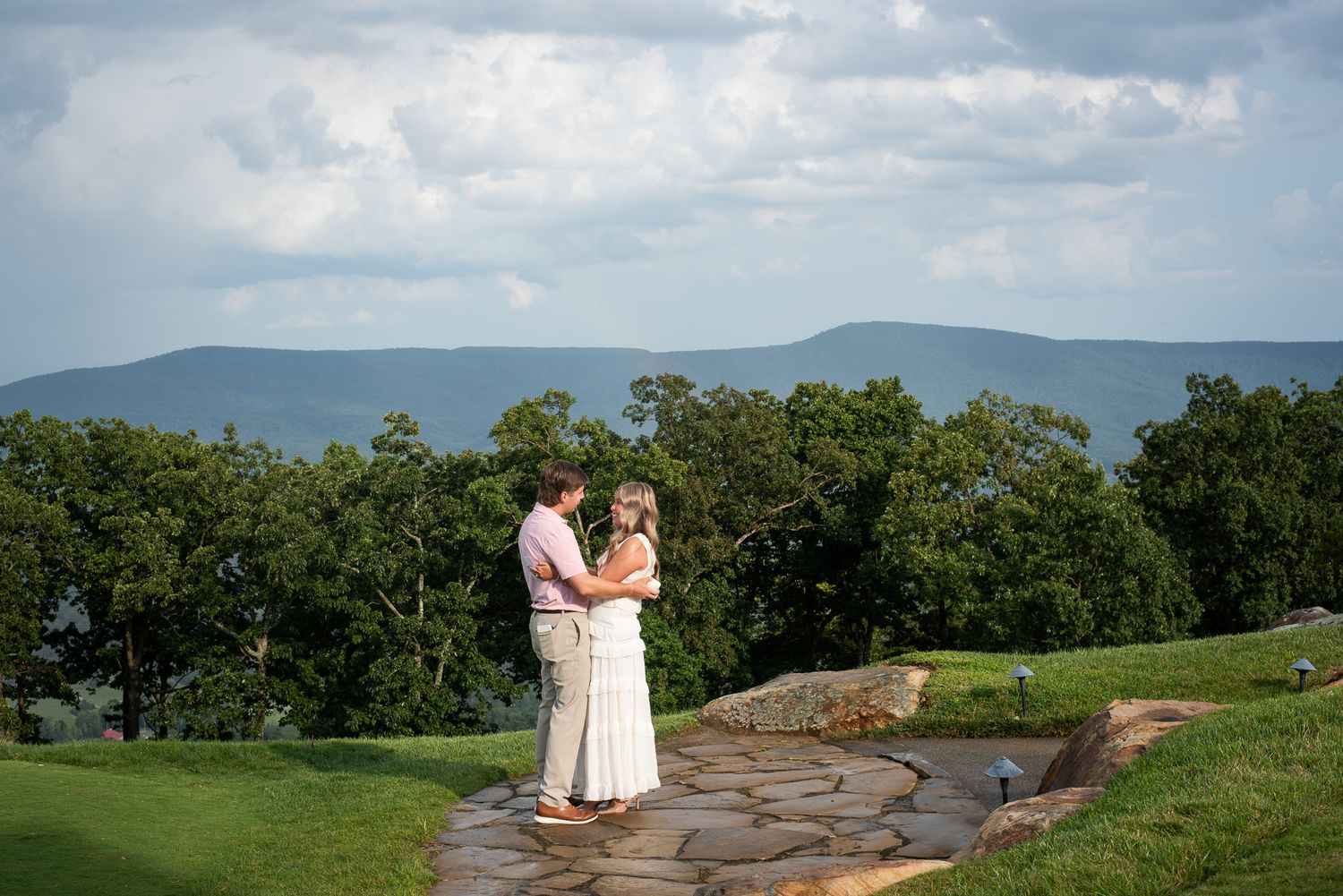 Couple standing face to face on stone path with stunning mountain landscape and dramatic sky behind.
