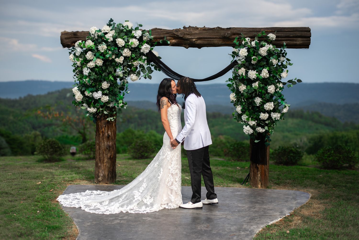 Bride and groom kissing under rustic wooden arch adorned with white floral arrangements outdoors.