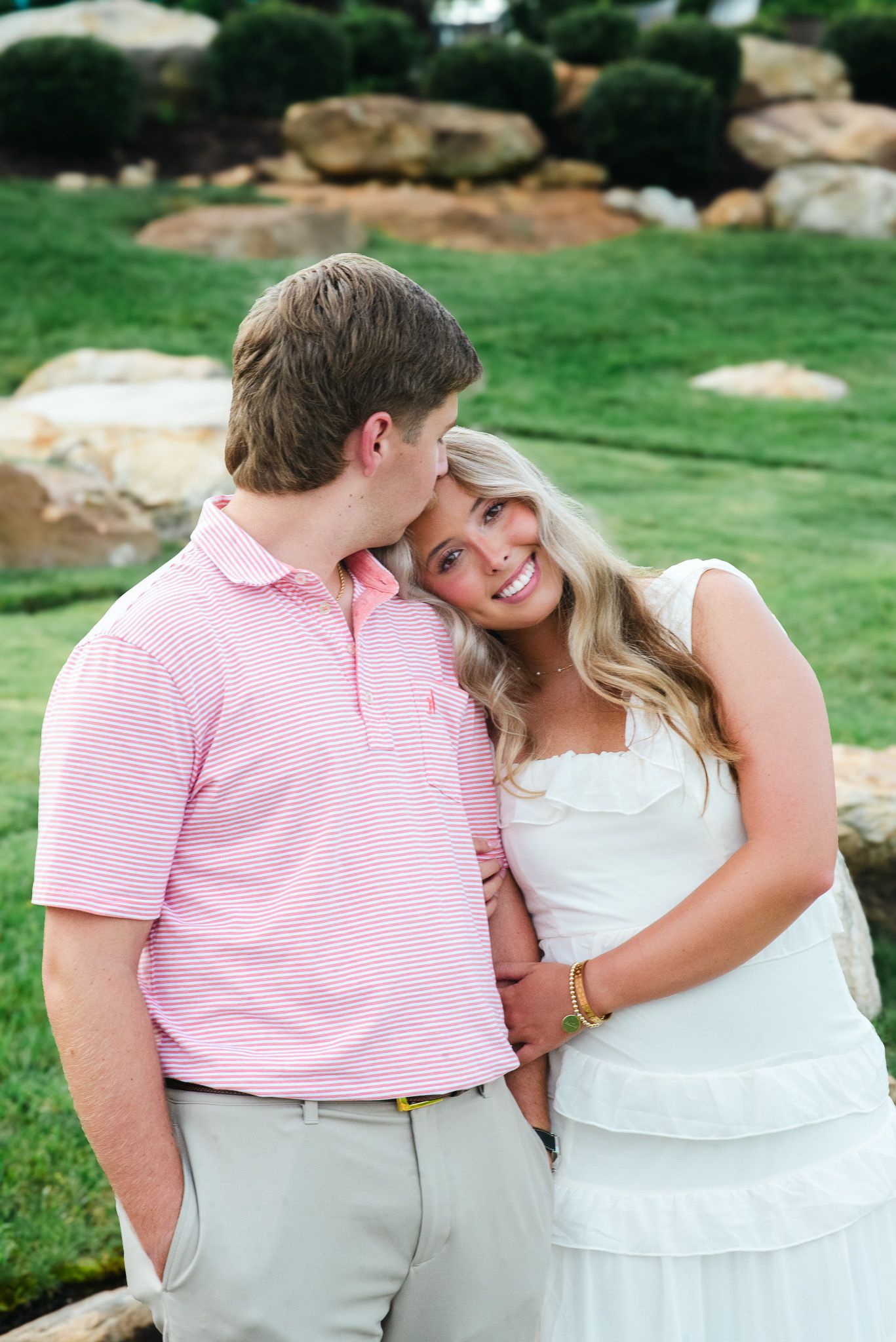 Couple posing near stone wall, man in pink striped polo kissing woman in white dress on cheek outdoors.