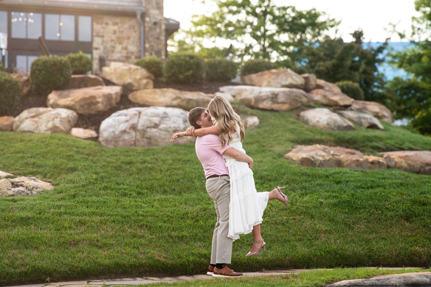 Romantic couple embrace as man lifts woman in white dress on scenic green lawn with stone backdrop.