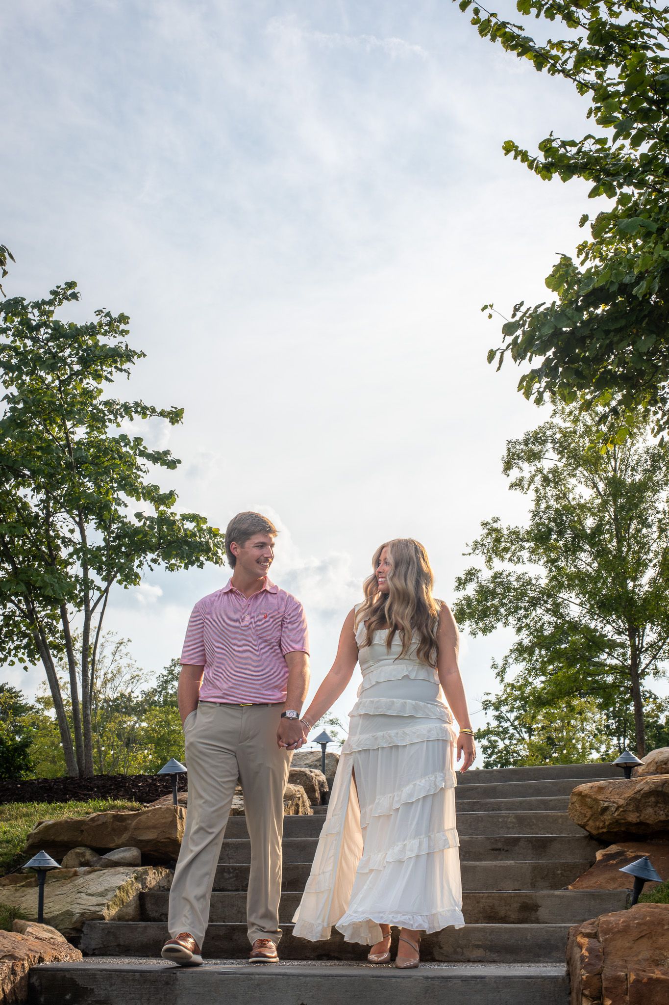 Engaged couple holding hands and smiling while walking down stone steps surrounded by lush green trees.