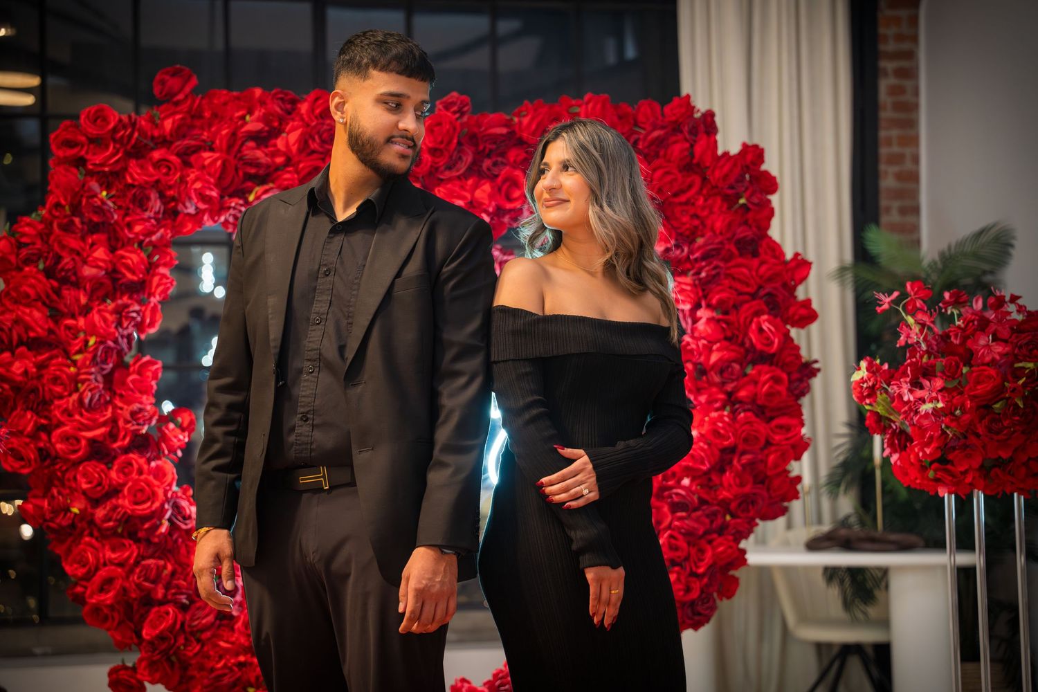 Stylish pair in black formal wear pose against an arch of red roses in romantic lighting.