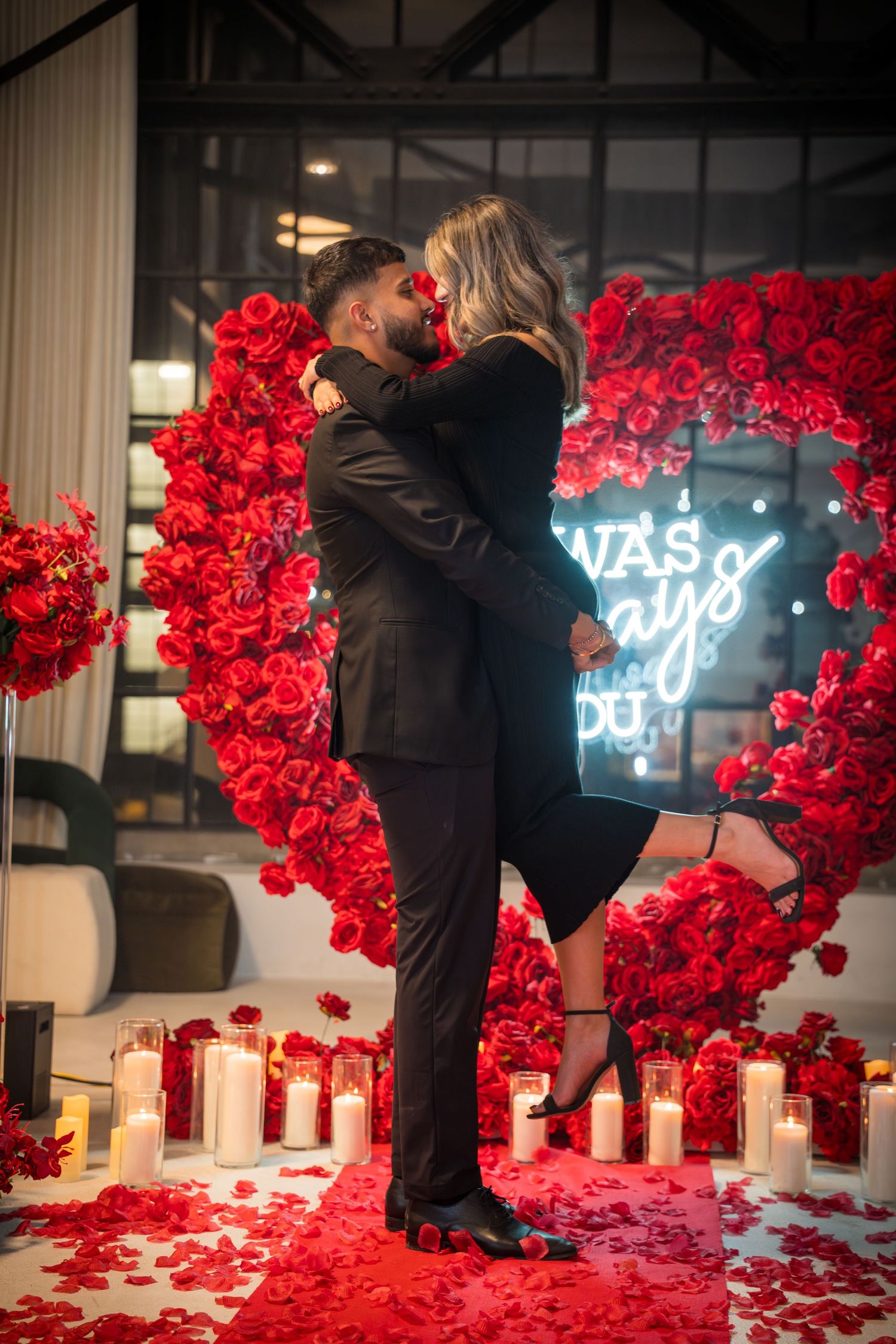 Romantic engagement scene with red rose arch, neon lights, and candles on floor.