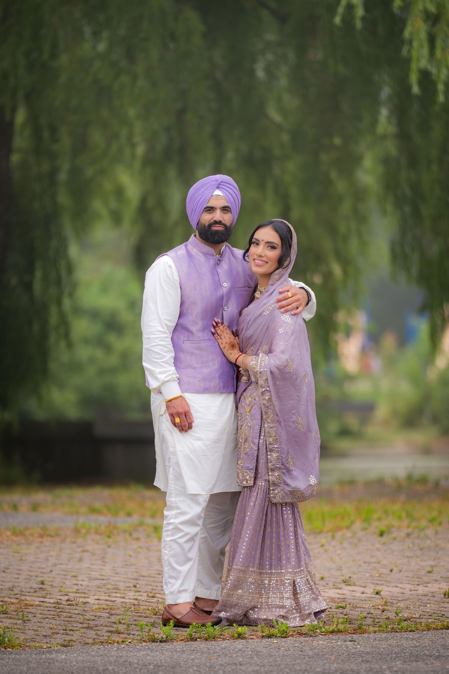 A couple in traditional Punjabi attire poses outdoors under willow trees wearing coordinated lavender and white outfits.