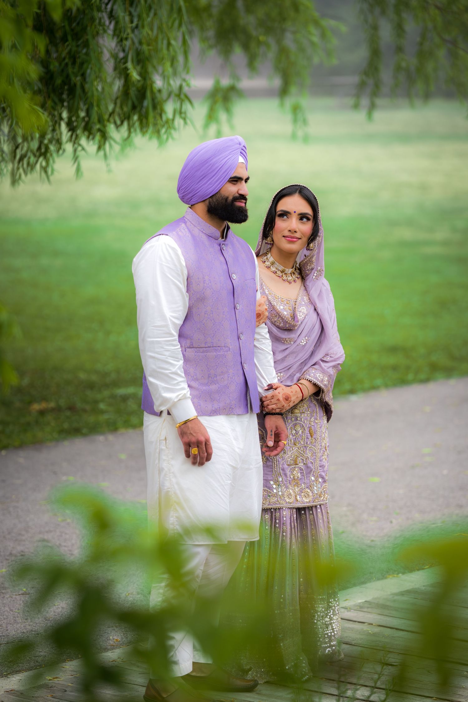 A couple in coordinated purple and white outfits photographed through willow branches in a garden.