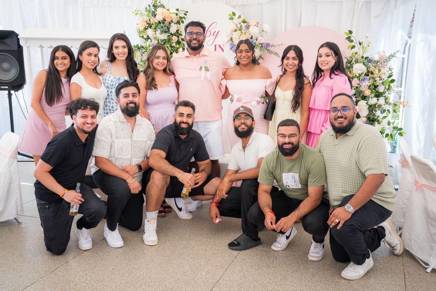 Large group photo of celebration guests in casual attire against floral backdrop.