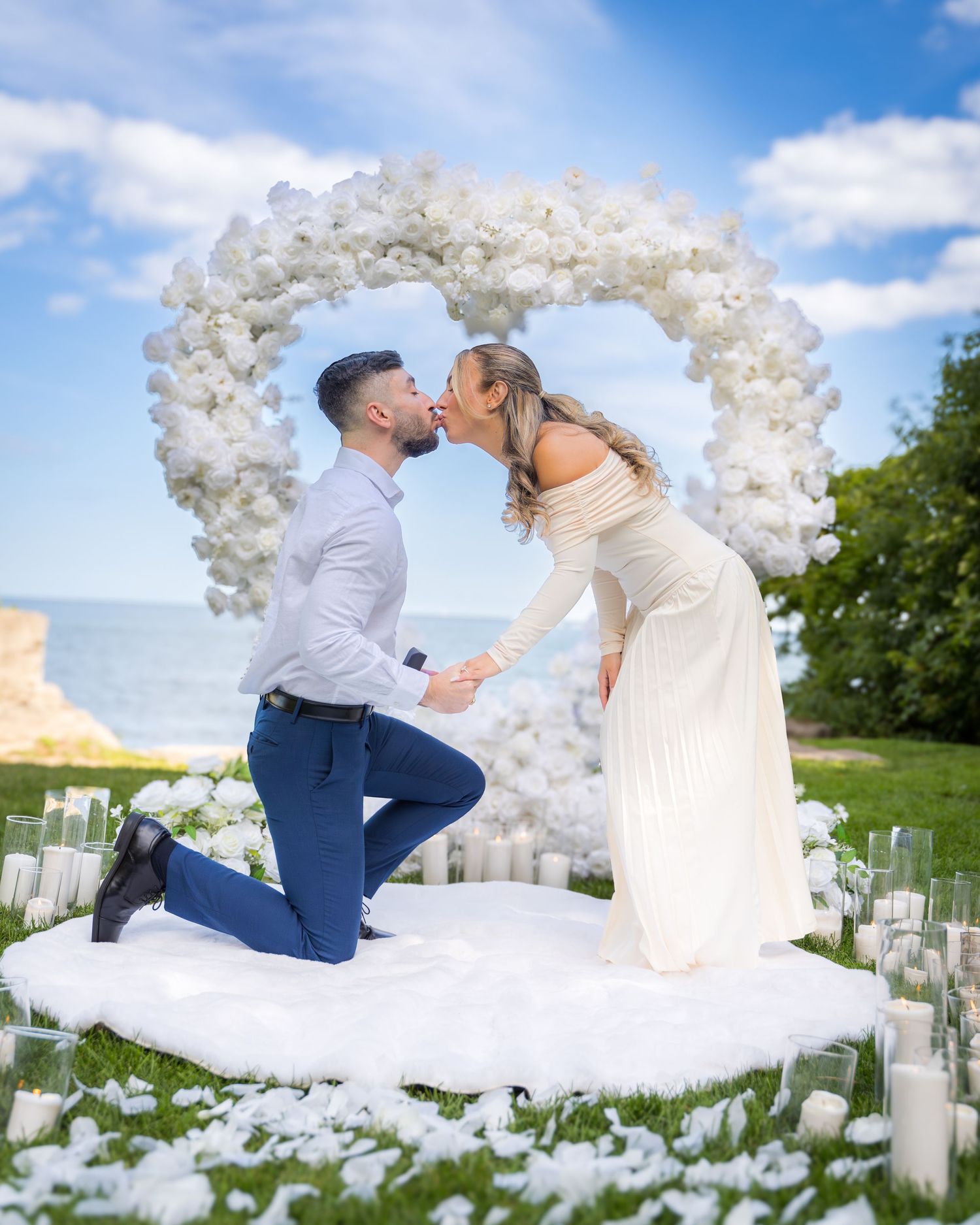 Beautiful proposal moment captured under a white floral arch with scattered candles and flowers.