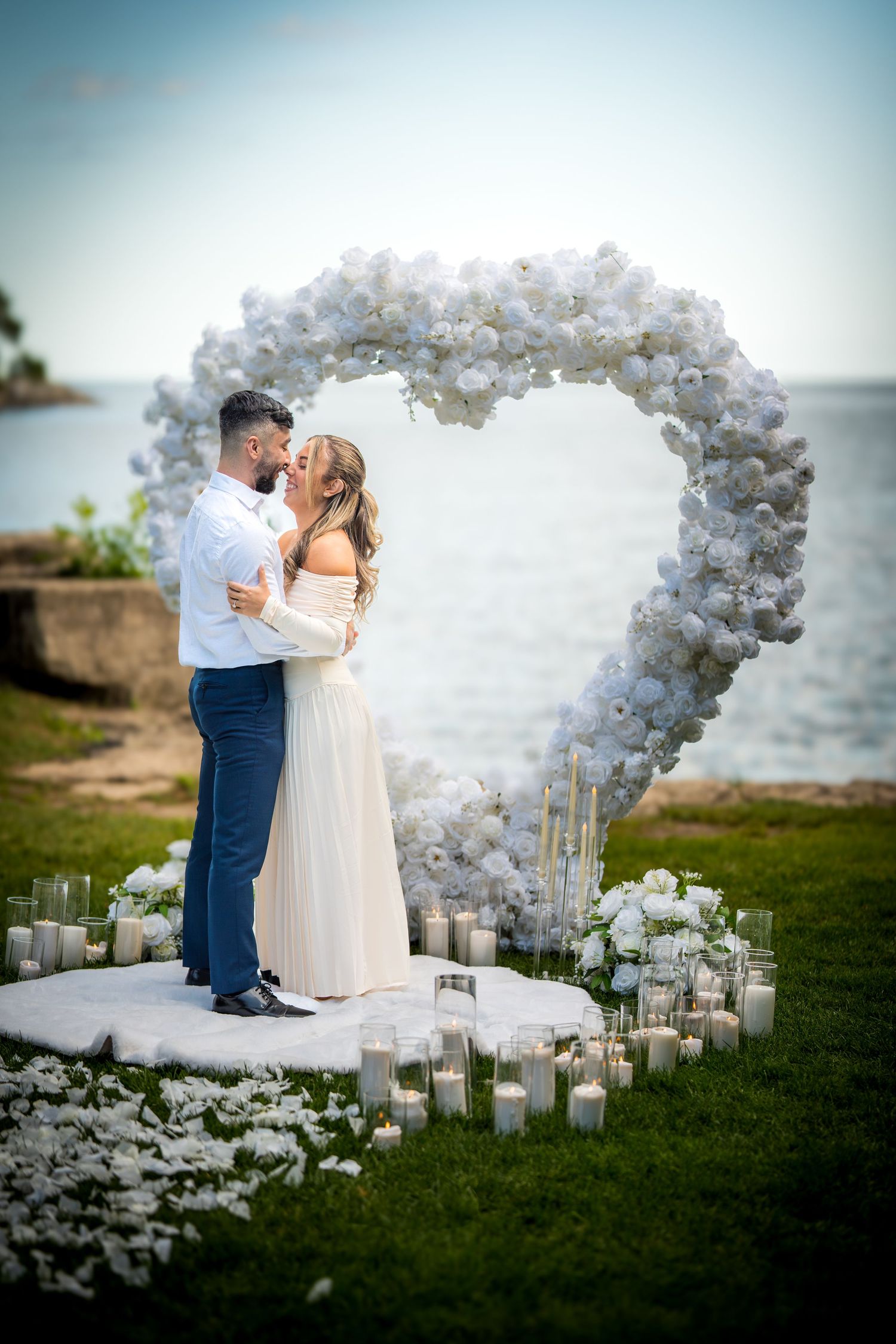 Couple embracing under a heart-shaped balloon arch with candles at oceanside proposal setting.
