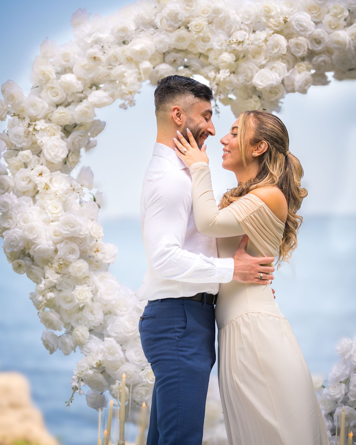 Wedding couple sharing tender moment beneath elaborate white flower arch installation.