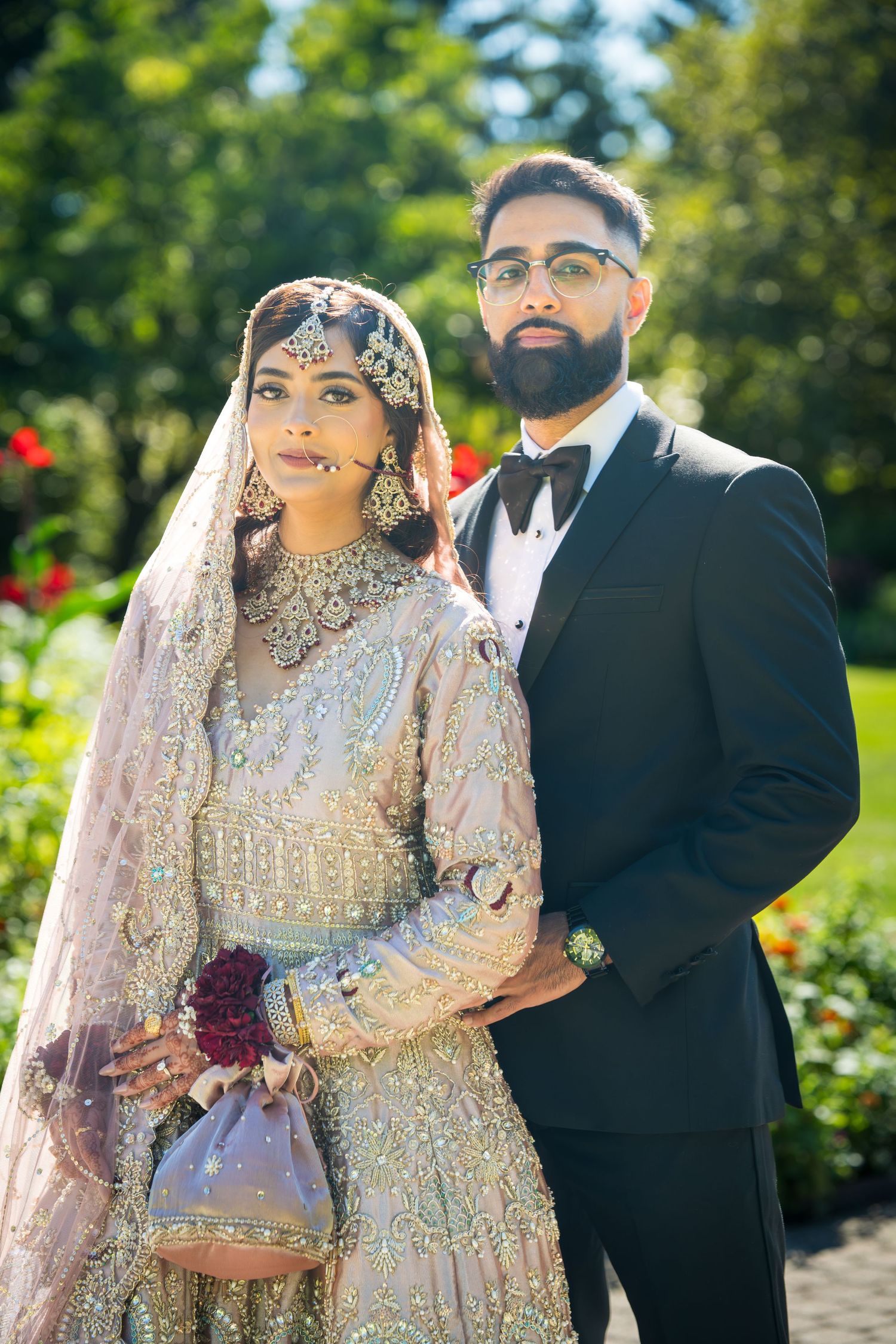 A couple in traditional South Asian wedding attire poses in a garden setting with vibrant flowers.