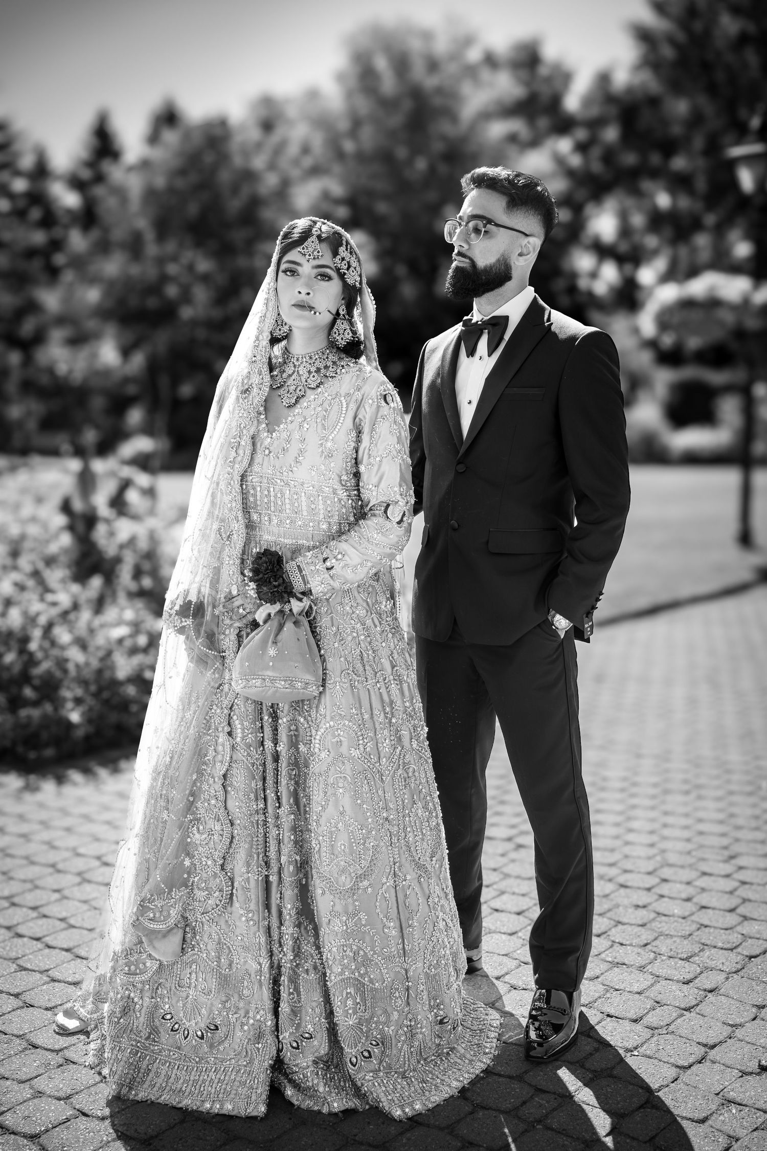 Black and white portrait of newlyweds in traditional South Asian wedding attire on a brick pathway.